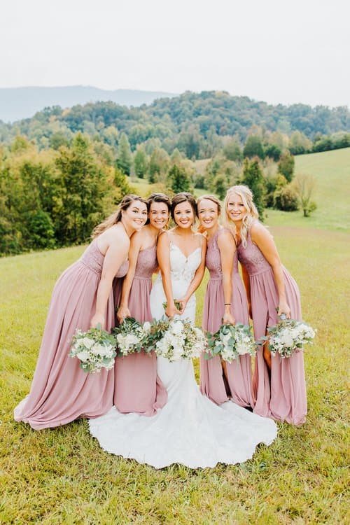 Bride and five bridesmaids in mauve dresses hold bouquets in a grassy field with rolling green hills. Moose Hollow Lodge.