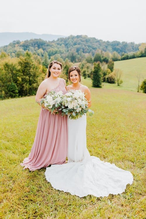 Bride in white dress and woman in pink dress holding bouquets in a grassy field with mountains. Moose Hollow Lodge venue.