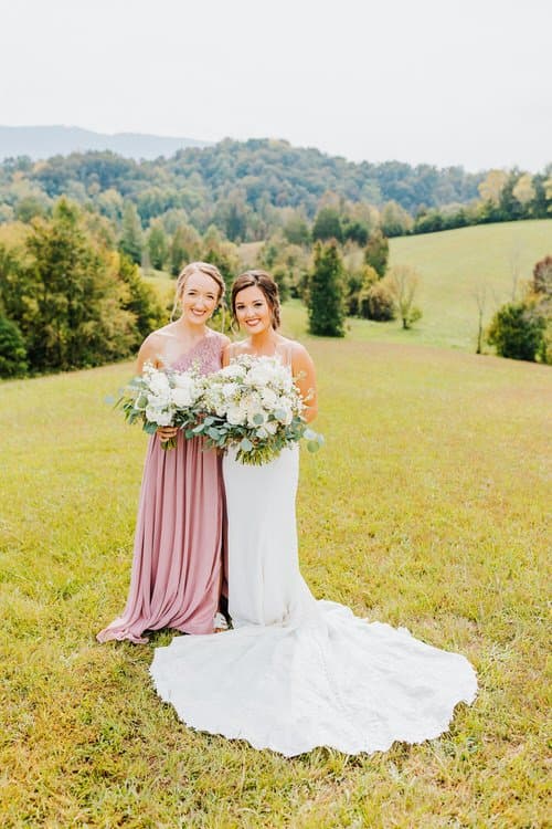 Bride in white gown and bridesmaid in pink dress hold bouquets in a grassy field with Smoky Mountain hills in background.