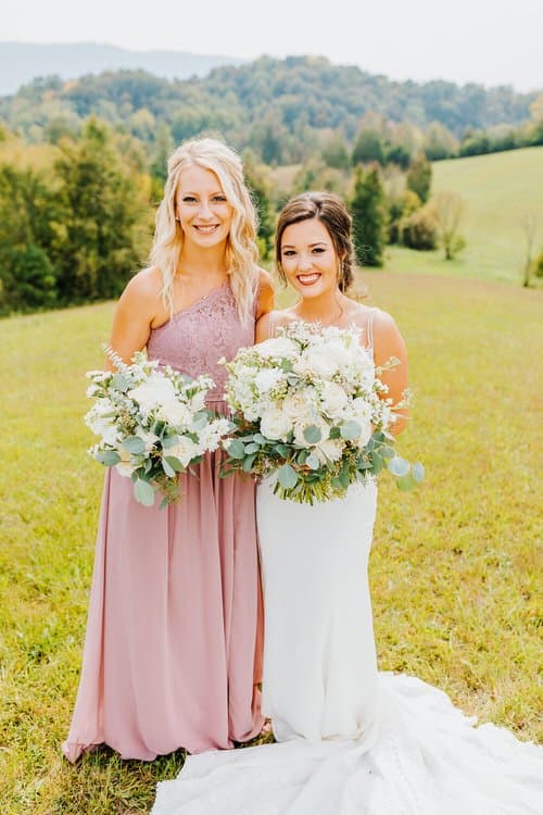 Bride in a white dress and bridesmaid in a pink dress hold white bouquets, standing in a grassy field with mountains.