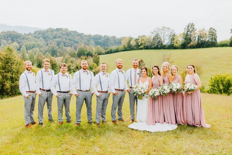 Wedding party poses in a field with rolling hills and trees. Groomsmen in grey pants, bride in white, bridesmaids in pink.
