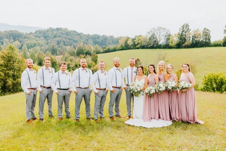 Wedding party in dusty rose and grey attire standing in a grassy field with rolling tree-covered hills in the background.