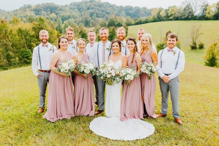 Wedding party with bride and groom, bridesmaids in dusty rose, groomsmen in grey and white, outdoors on a grassy hill...