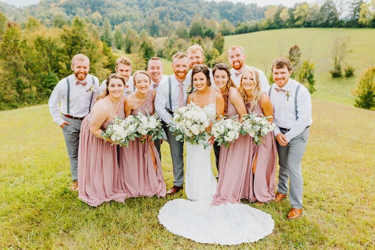 Wedding party of 12 smiling outdoors: bride, five bridesmaids in mauve dresses, six groomsmen. Green hills and trees...