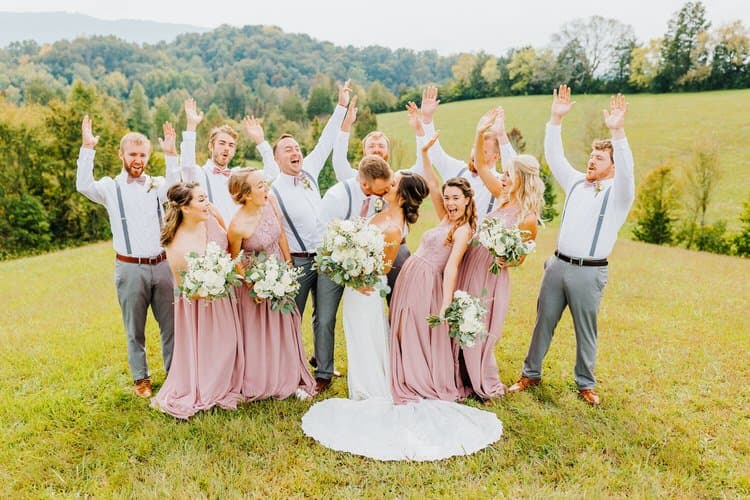 Wedding party, bride and groom kissing, bridesmaids in pink, groomsmen in grey, on a grassy hill with trees and mountains.