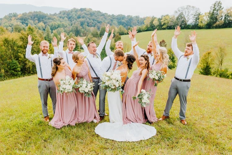 Wedding party: bride, groom, five bridesmaids, five groomsmen in a grassy field, forested mountains behind. Couple kisses.