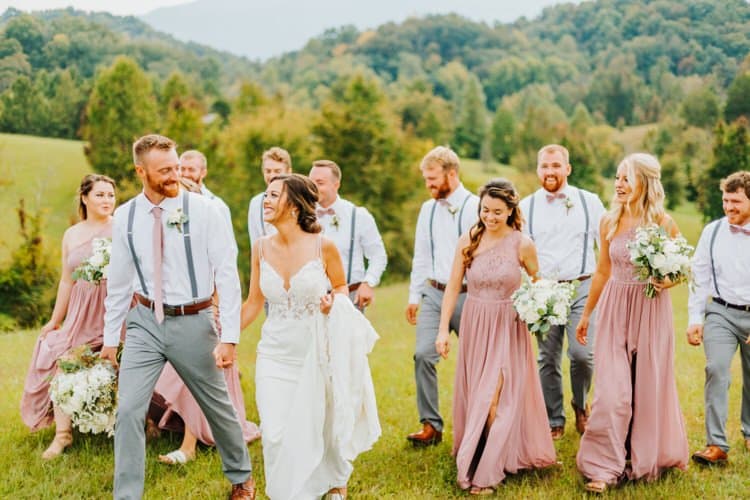 Wedding party walks through a grassy field with wooded hills and mountains in the background at Moose Hollow Lodge.