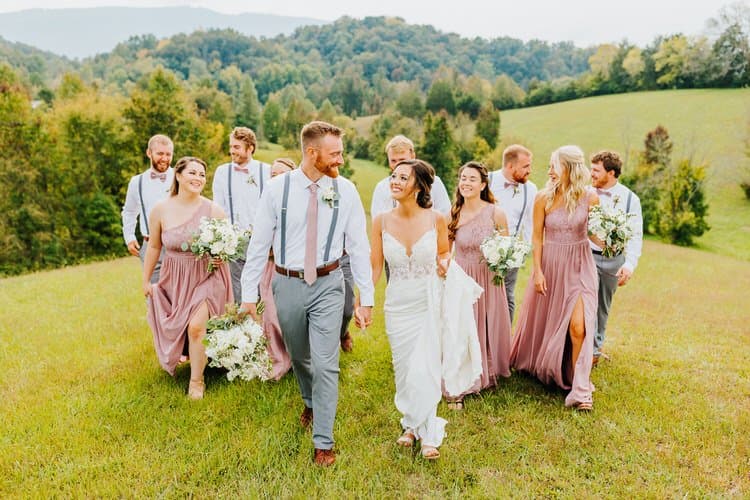 Wedding party walks down a grassy field with rolling hills and trees in the background. Bride, groom, bridesmaids, and...