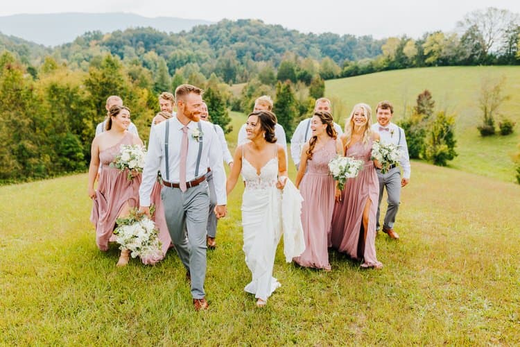 Wedding party, including bride and groom, walking up a grassy hill with rolling forested hills in the background.
