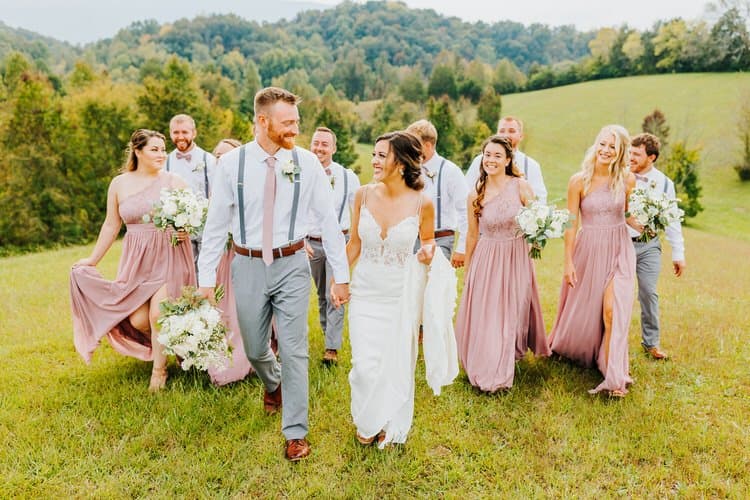 Bride and groom hold hands walking with wedding party in a grassy field with wooded hills in background.