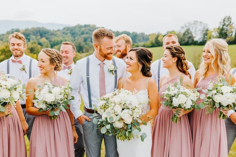 Wedding party with a bride, groom, bridesmaids, and groomsmen posing outdoors. Rolling green hills and trees are in the...