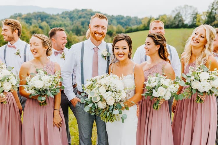 Wedding party with bride, groom, bridesmaids in rose dresses, groomsmen in suspenders, standing on grassy hill.