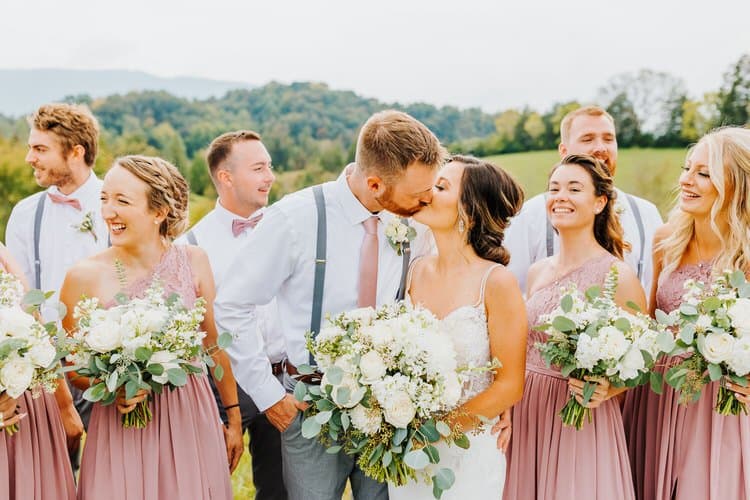 Bride and groom kiss with their wedding party in an outdoor setting, surrounded by green hills and trees under a light sky.