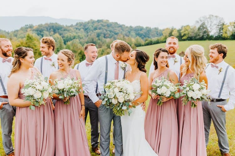 Wedding party poses in a grassy field with a mountain background. Bride and groom kiss, surrounded by guests.