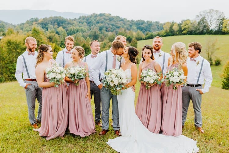 Wedding party in a field, bride and groom kissing, bridesmaids in dusty rose, groomsmen in grey pants, mountain backdrop.