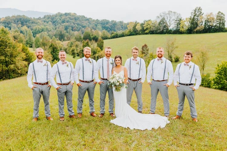 Bride with six groomsmen in a grassy field with rolling hills and trees. Outdoor wedding party photo at Moose Hollow Lodge.