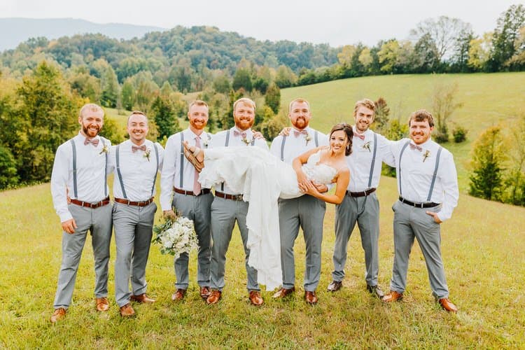 Bride carried by groomsmen in a field with rolling green hills. Groomsmen wear grey pants, white shirts, bow ties.