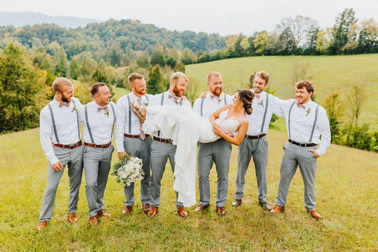Bride carried by groomsmen in a grassy field at Moose Hollow Lodge with a mountain backdrop under an overcast sky.
