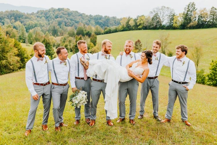 Bride in white dress held by groom and six groomsmen in gray pants, white shirts, and suspenders in a grassy field with...