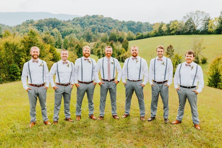 Seven groomsmen in white shirts, gray pants, and suspenders stand in a grassy field with rolling hills and trees.