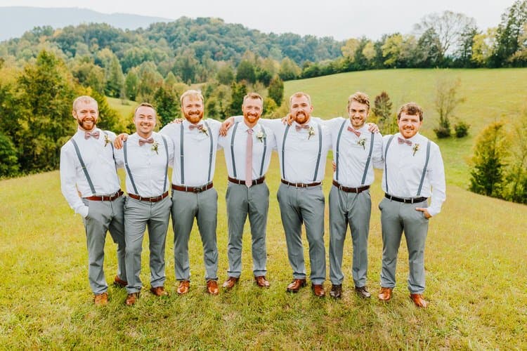 Seven men in white shirts, grey pants, pink ties, and suspenders pose on a grassy hill with tree-covered mountains.