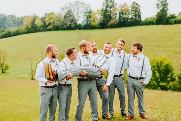 Seven men in white shirts, grey pants, and suspenders pose in a grassy field with trees. One man is carried horizontally.