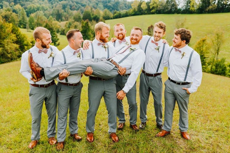 Seven men in white shirts, grey pants, and pink bow ties in a grassy field, with one man being carried by two others....