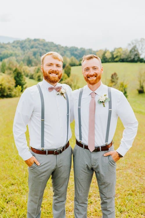 Two men in white shirts, grey suspenders, grey pants, and pink accessories stand in a grassy field with green hills.