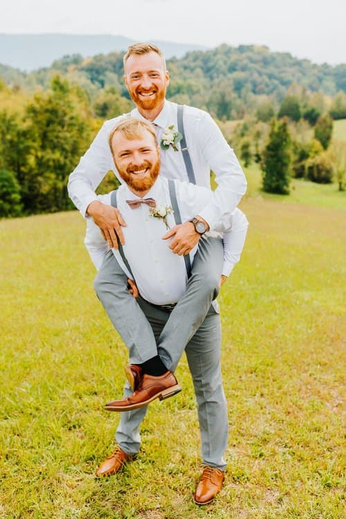 Two men in white shirts, grey pants, and suspenders, one giving a piggyback ride, smile outdoors in a grassy field with...