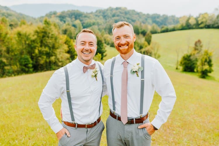 Two men in white shirts, grey suspenders, and pink accessories pose in a grassy field with rolling green hills and trees.