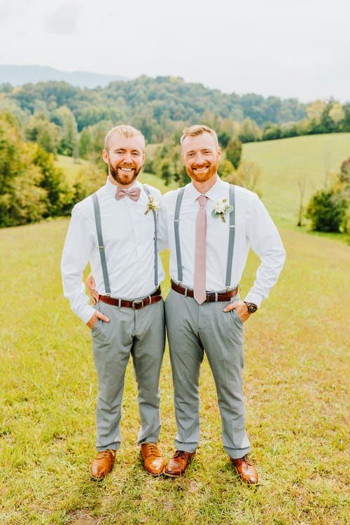 Two men in white shirts, grey pants, suspenders, and pink ties stand in a grassy field with wooded hills.