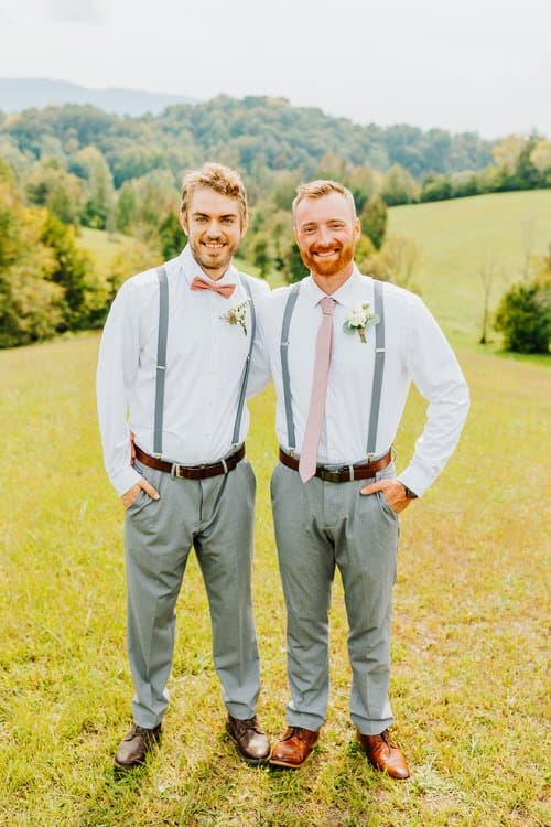 Two groomsmen in suspenders, white shirts, and grey pants stand in a grassy field with hills at Moose Hollow Lodge.