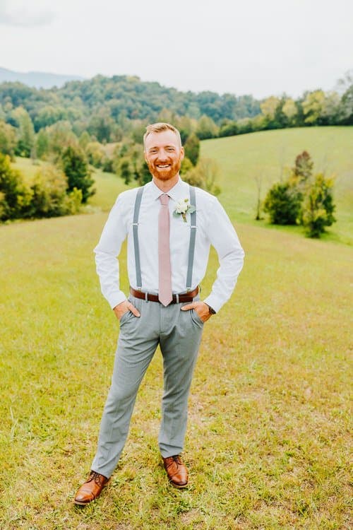 Man in white shirt, grey suspenders, blush tie, and grey pants with boutonniere stands in a grassy field. Hills and trees...