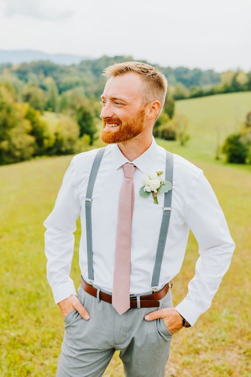 Man in white shirt, pink tie, and gray suspenders and pants, standing outdoors with green rolling hills and mountains...