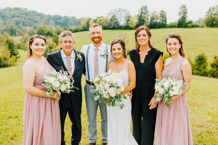 Wedding group portrait in an open grassy field with distant green hills. Bride, groom, two bridesmaids, and two adults.