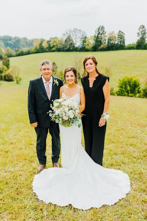 Bride in white gown with father and mother in grassy field. Rolling hills and trees in background. Outdoor family photo.
