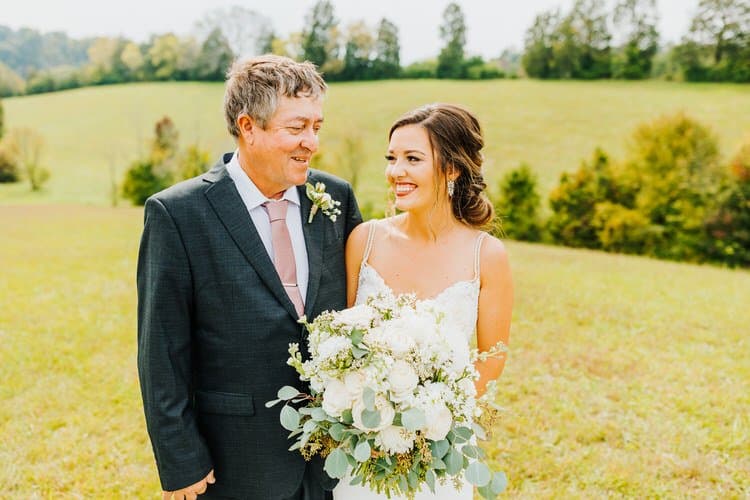 Bride and man in suit, holding white bouquet, stand in a sunny grassy field with distant trees on a wedding day.
