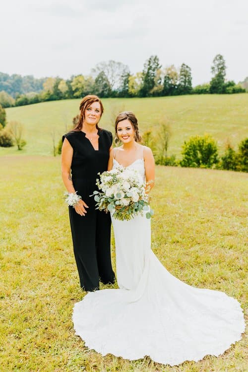 Woman in white wedding dress holding bouquet stands beside woman in black jumpsuit with corsage in a grassy field. Hills...