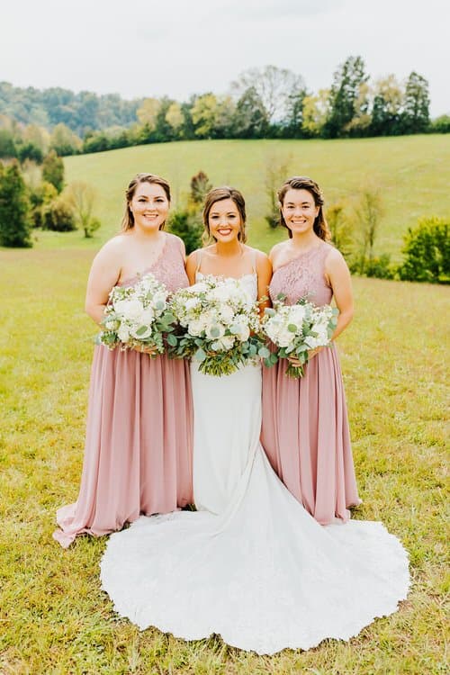 Bride in white gown with train and two bridesmaids in dusty rose dresses hold bouquets in a grassy field with hills.