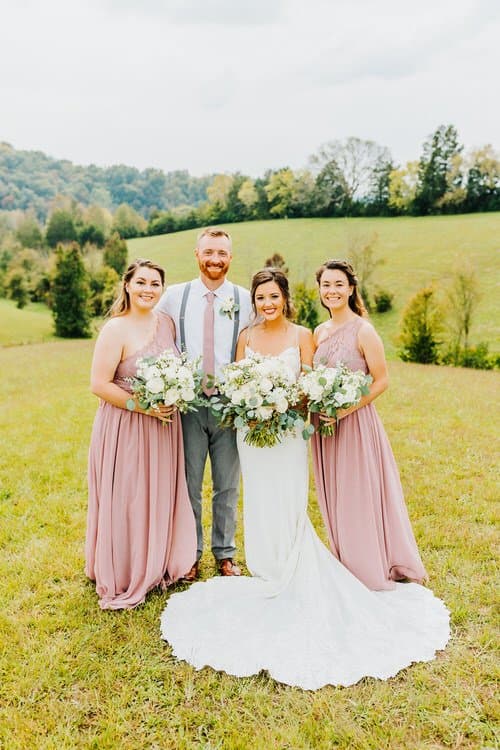 Wedding party of four: bride, groom, two bridesmaids. They stand in a grassy field with green hills and trees in the...