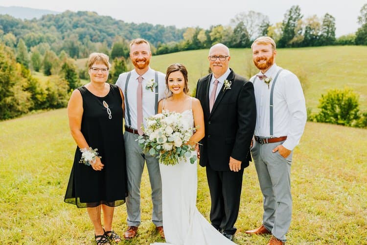 Bride with a white and green bouquet, four family members stand in a grassy field with tree-covered hills.
