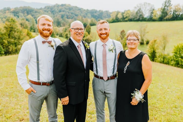 Four adults, dressed in wedding attire, stand in a sunny green field with distant tree-covered mountains.