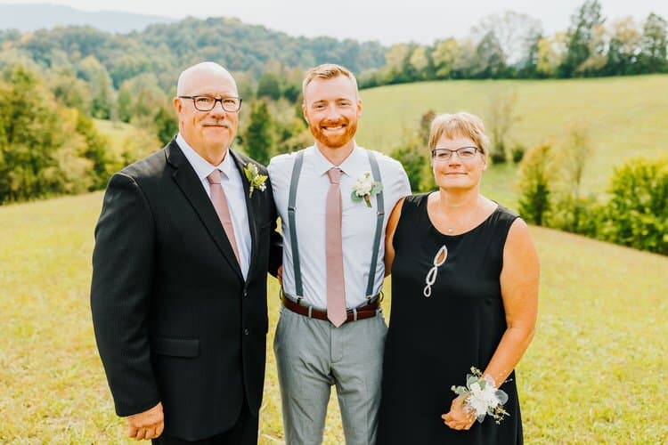Three adults, two men and one woman, in wedding attire pose in a grassy field with green mountains in the background.