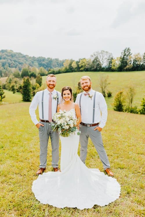 Bride in a white gown with a bouquet stands with two men in white shirts, grey pants, and suspenders in a field.