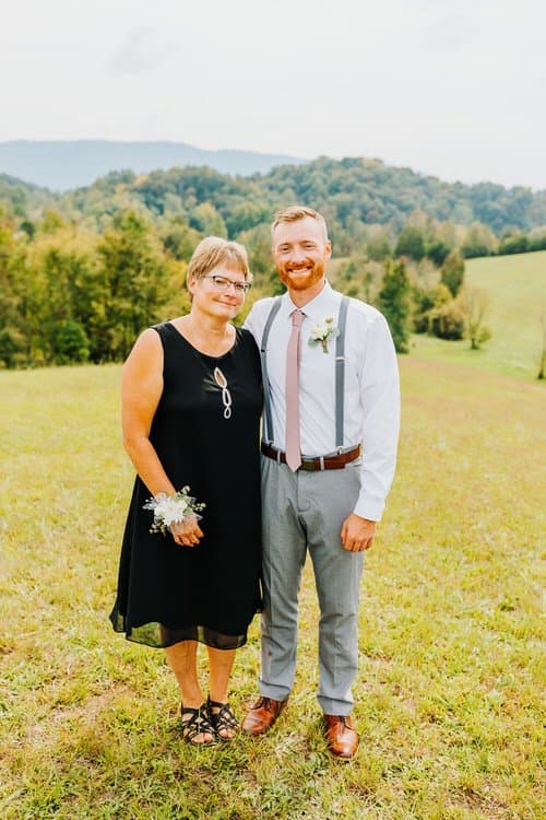 Man in shirt, tie, suspenders, boutonniere with woman in black dress, corsage. Standing in field with hills.