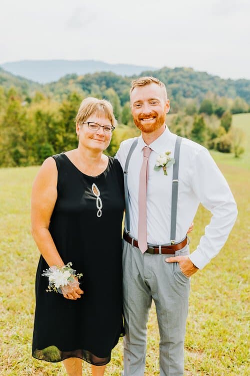 Woman in black dress and corsage, man in white shirt, pink tie, and suspenders in a grassy field with distant mountains.