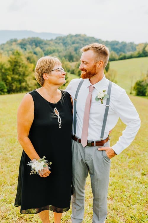 Woman in black dress with wrist corsage and man in white shirt, tie, and suspenders in grassy field with mountains. Moose...