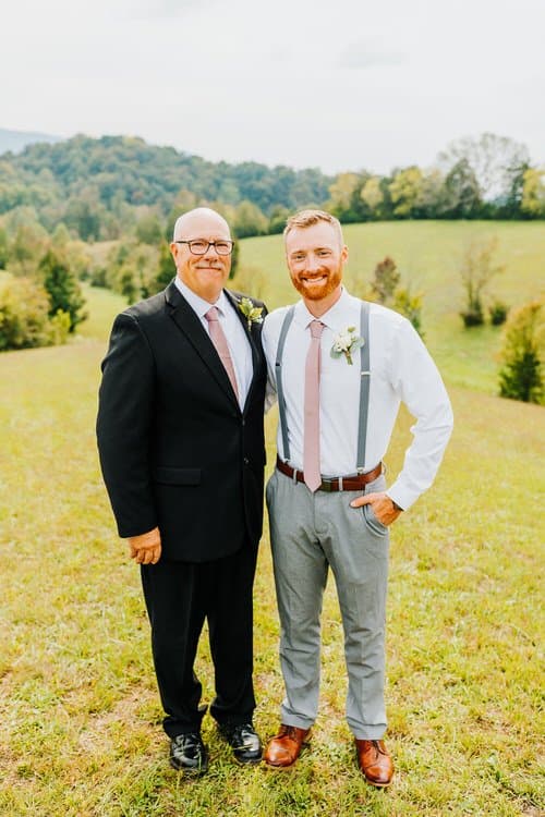 Two men in wedding attire, one in a suit, one in suspenders, stand in a field with distant tree-covered hills.
