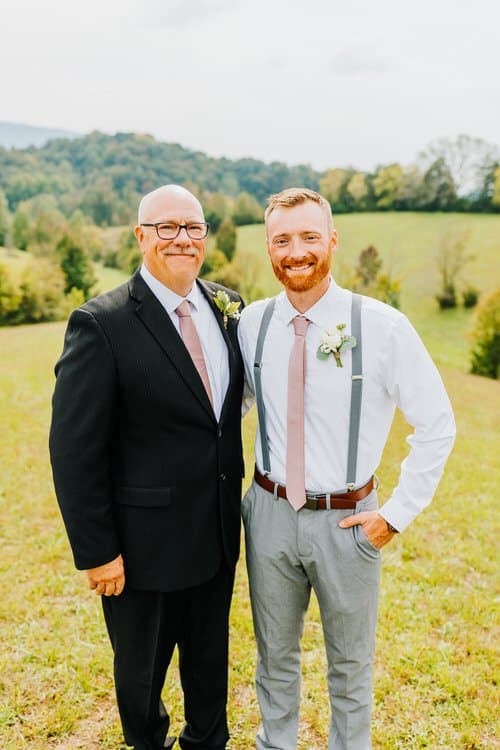Two men in wedding attire, one in a suit and one in suspenders, stand in a grassy field with mountains at Moose Hollow Lodge.