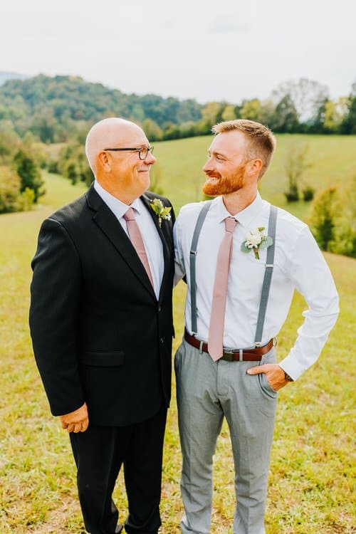 Two men in wedding attire stand in a grassy field, looking at each other. Rolling green hills in the background.